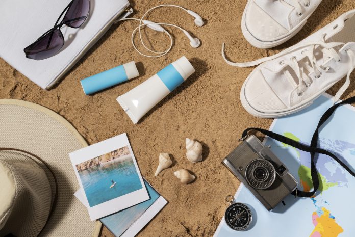Flat lay of essential beachwear and travel items including sunglasses, hat, swimwear, sandals, and travel accessories arranged for a beach vacation.